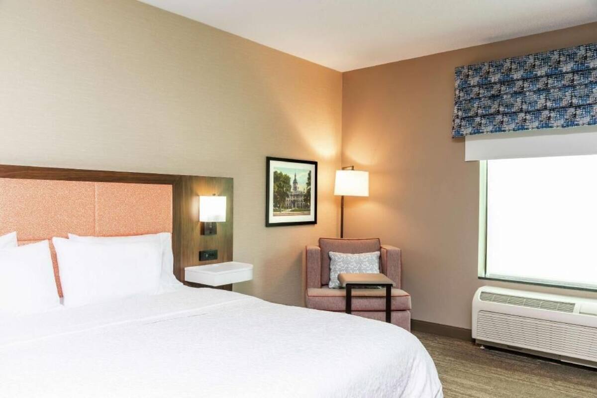 A hotel room with a large bed, a small table, a chair, a nightstand, a lamp, a picture on the wall, and an air conditioning unit.