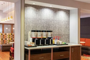 Uma estação de café moderna com vários dispensadores, uma pequena mesa com chávenas e acompanhamentos, e um espaço de estar com uma parede colorida e um sofá.