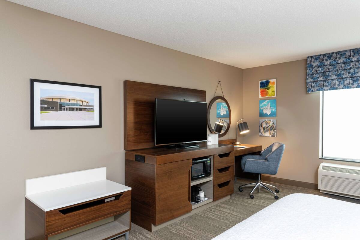 A hotel room with a bed, a desk, a television, a microwave, a chair, and artwork on the walls.