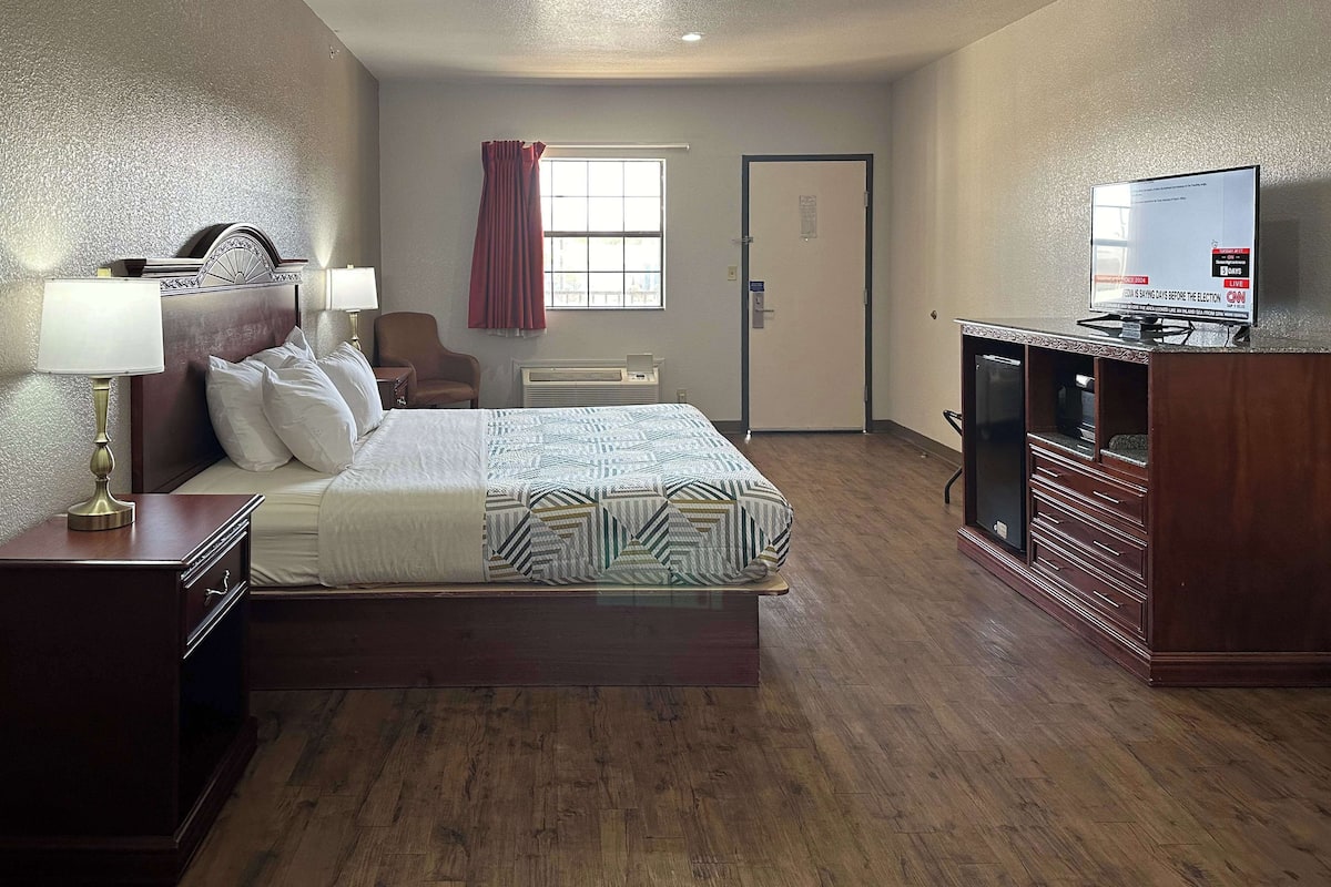 A hotel room with a bed, bedside table, lamp, chair, TV, and a door.