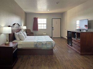 A hotel room with a bed, bedside table, lamp, chair, TV, and a door.