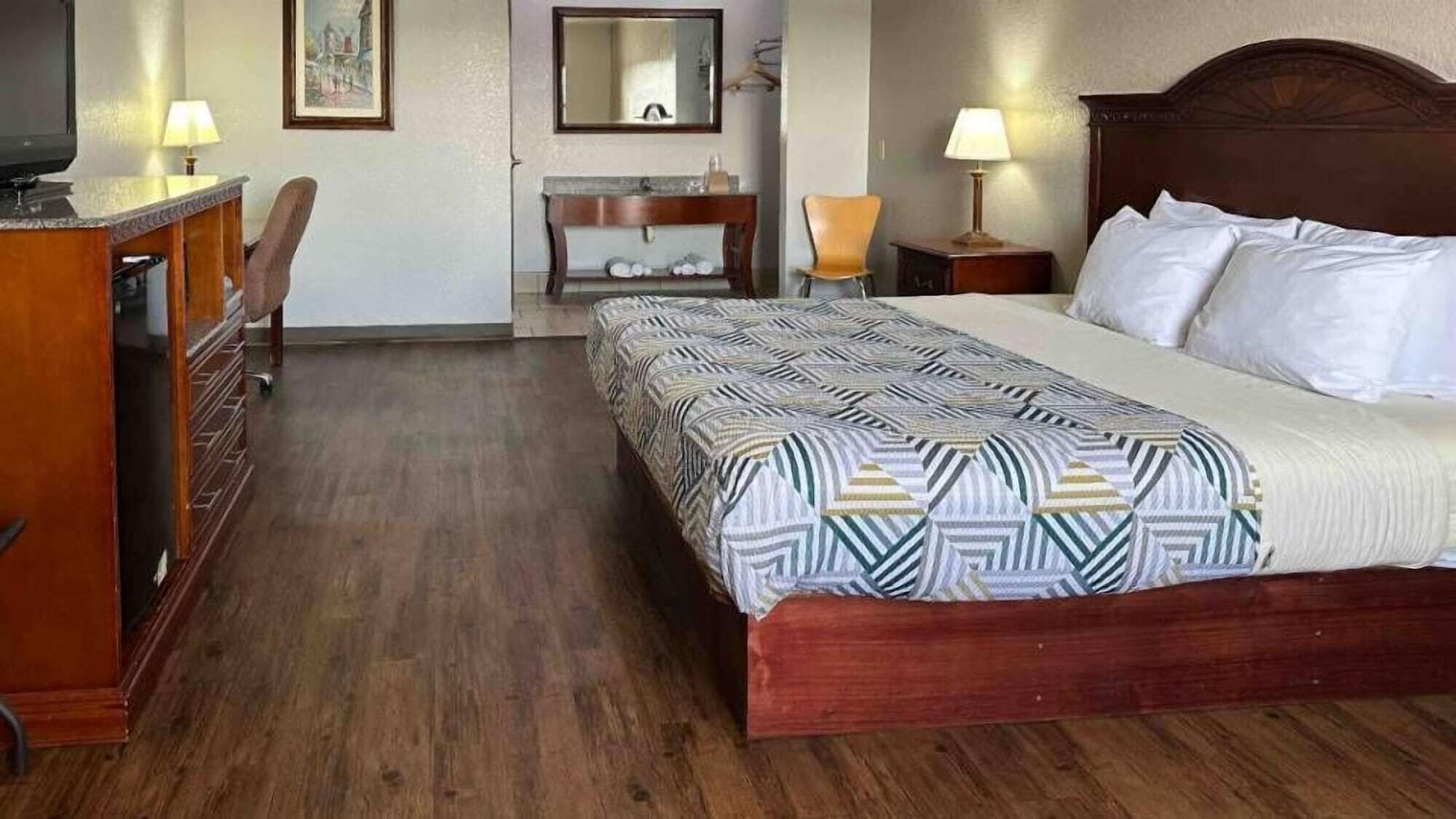 A hotel room with a wooden bed, a desk with a chair, a dresser, a mirror, and a lamp.