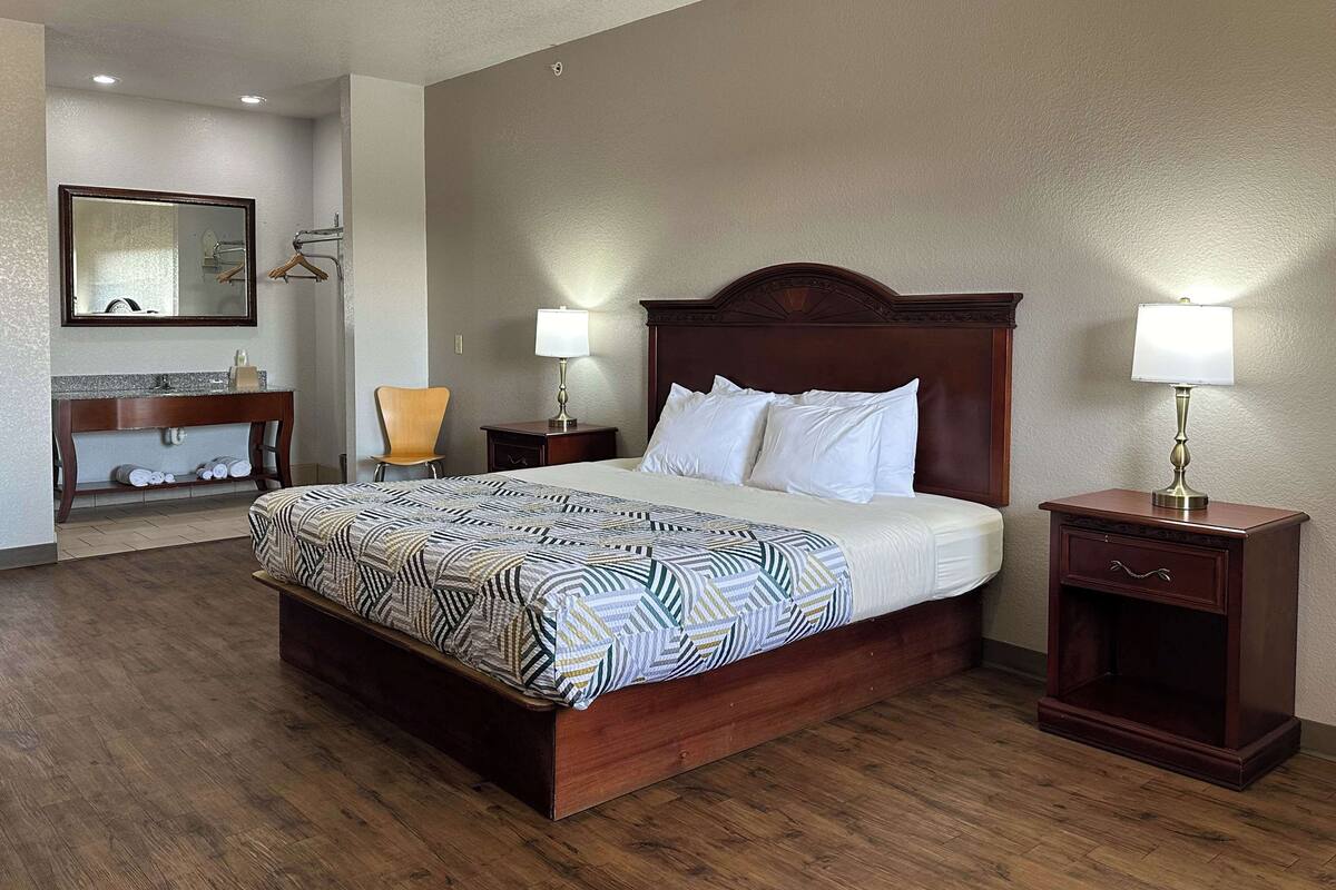 A hotel room with a large bed, two bedside tables with lamps, a wooden nightstand, a wooden chair, a bathroom with a mirror, and a towel rack.