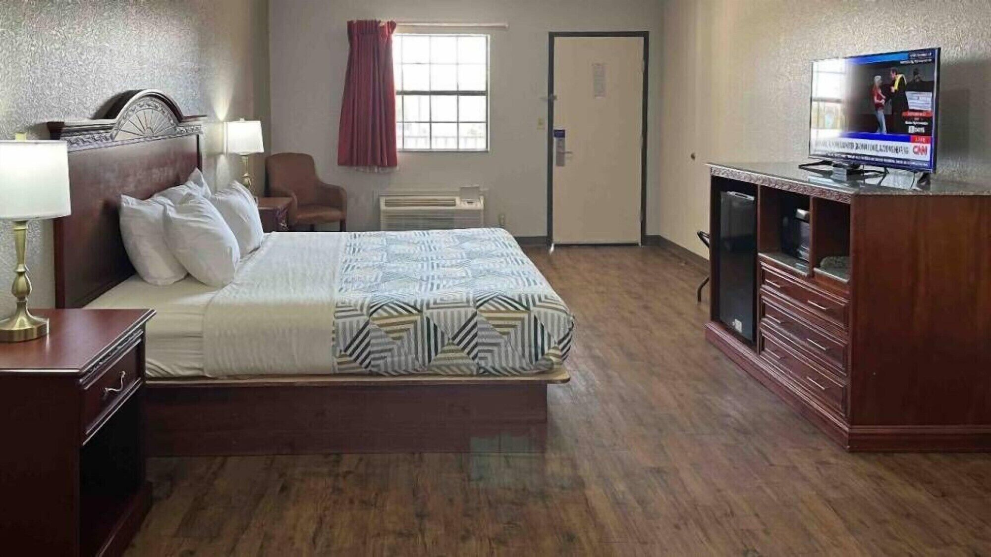 Quarto de hotel com cama, escrivaninha com luminária, televisão, cadeira e janela com cortinas.