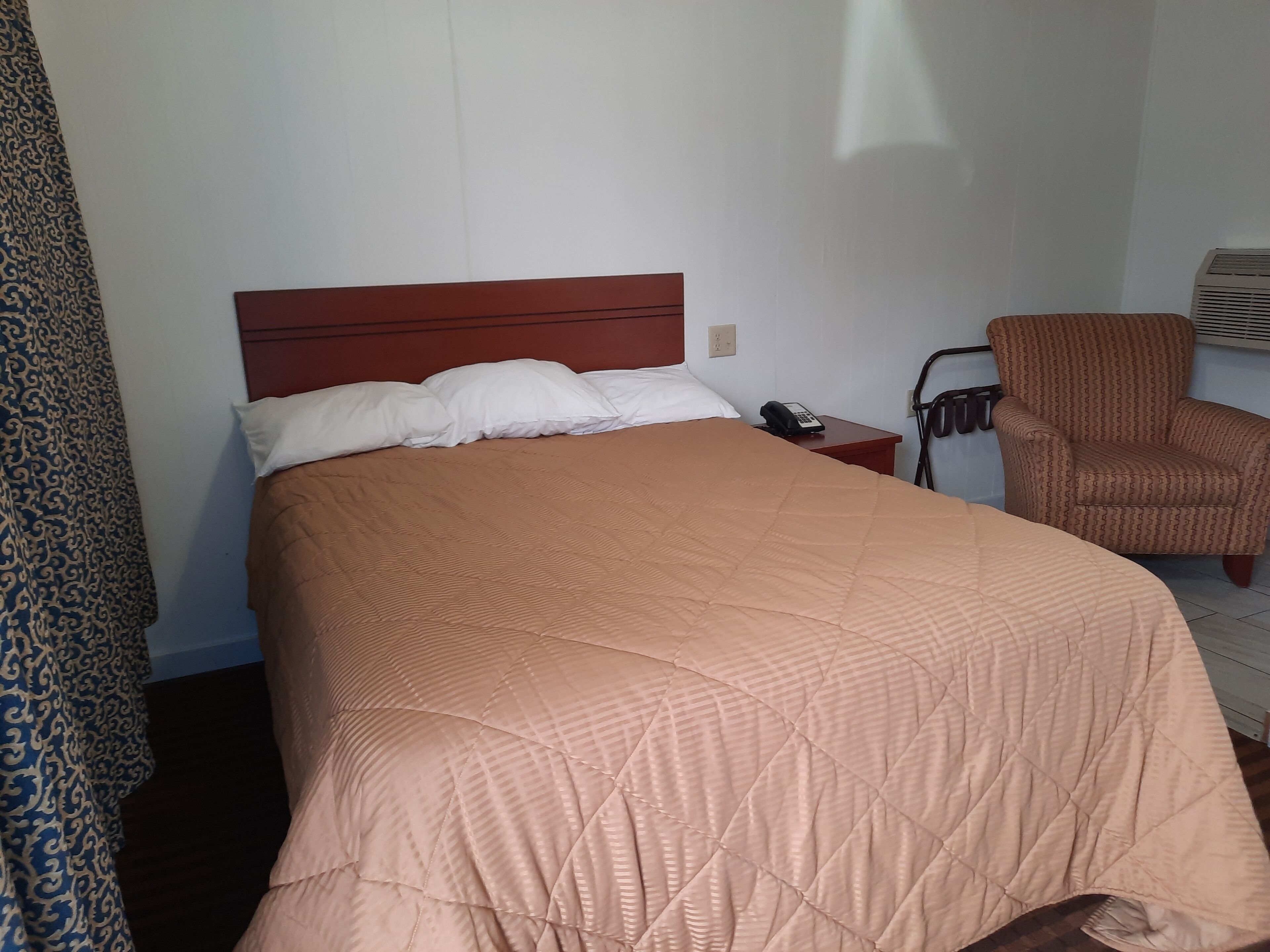 A hotel room with a bed, a chair, a telephone, and a wall-mounted air conditioning unit.