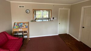 A room with a red couch, a small window with a mirror, a white door, and a shelf with books. - Motel Reedsburg (Reedsburg)