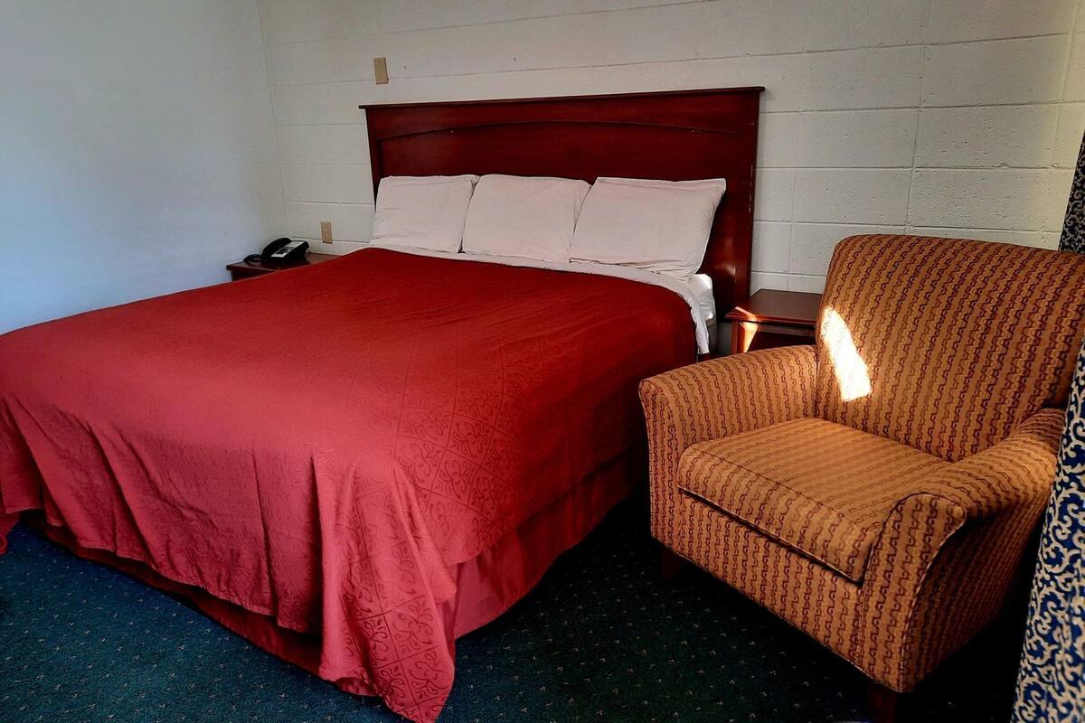 A single bed with a wooden headboard, a red bedspread, and a patterned armchair in a room with white walls and a blue carpet.
