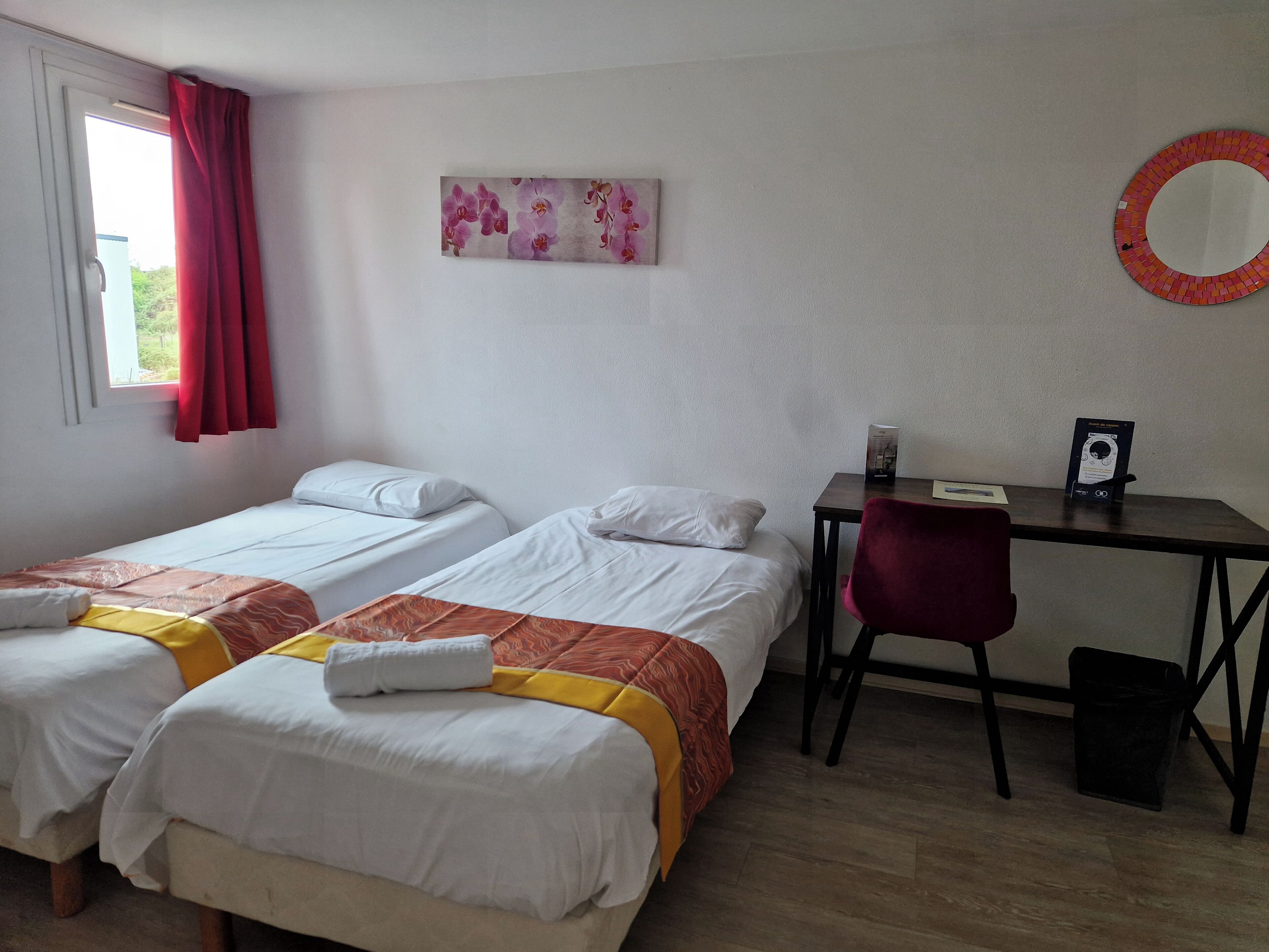 A room with two single beds, a desk, a chair, a mirror, and a window with curtains.