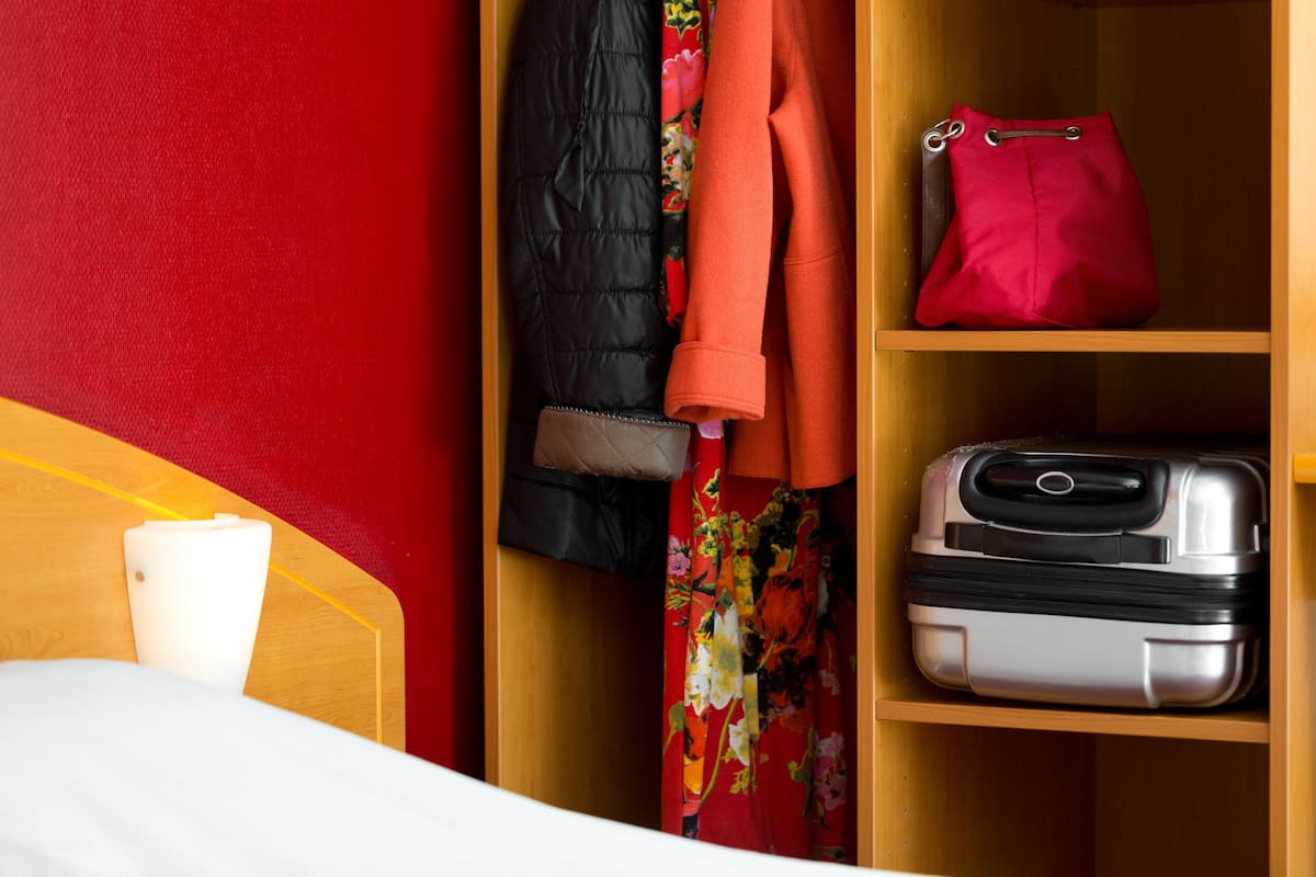 Una cama, un armario de madera con una pared roja, una chaqueta negra, un abrigo naranja, un bolso rojo y una maleta.