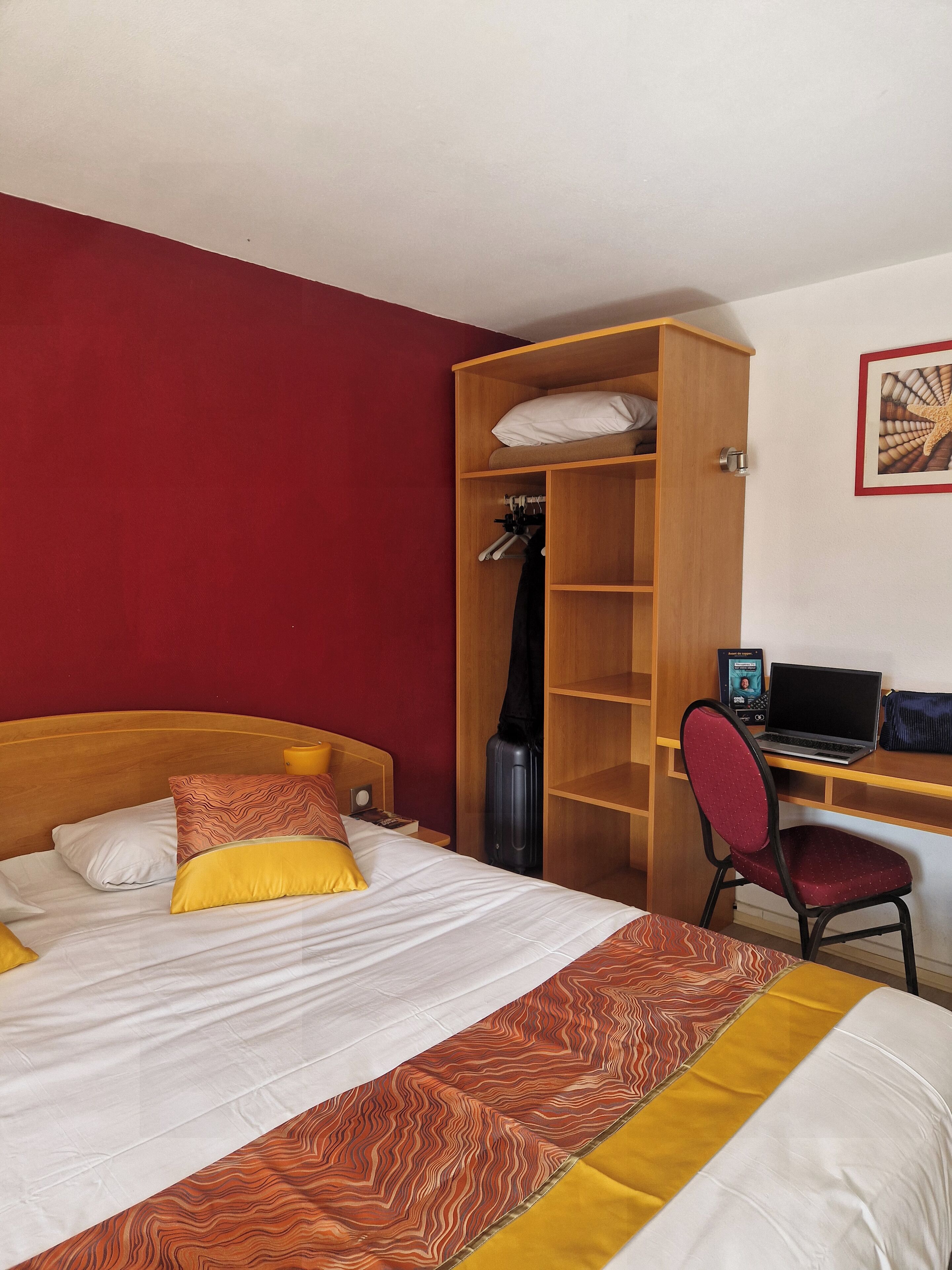 A hotel room with a bed, a desk with a laptop, a chair, a wardrobe, and a guitar.
