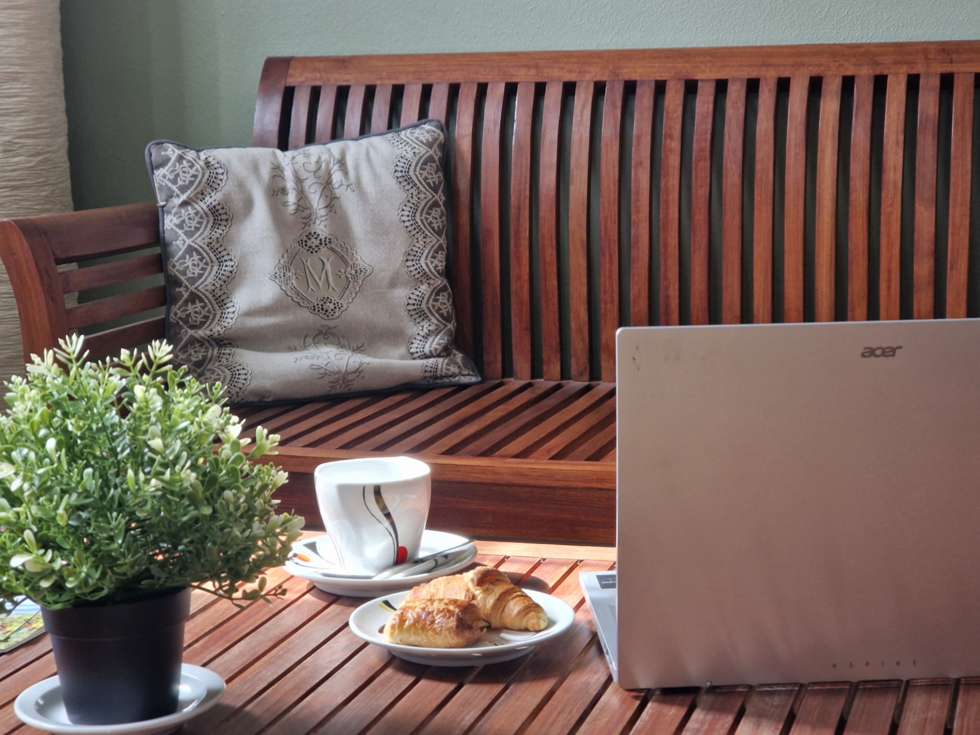 Ein Holztisch mit einem Laptop, einer Tasse, einem Untertasse, einem Teller mit Gebäck und einer Topfpflanze.
