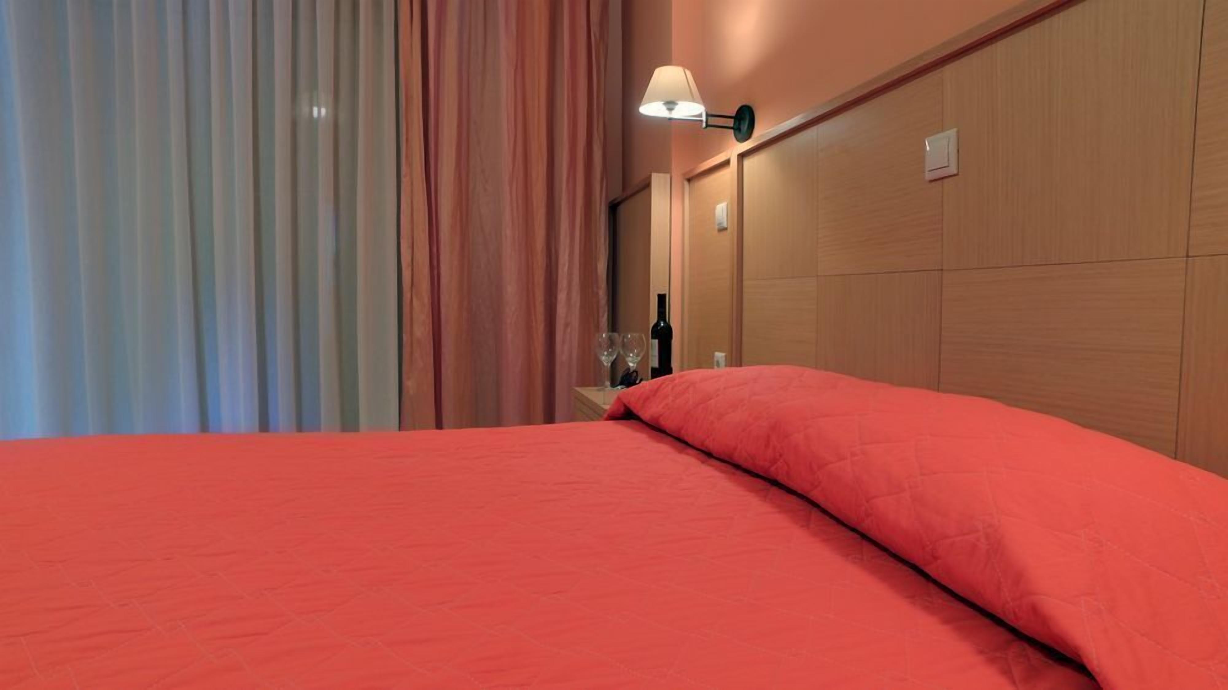 Habitación de hotel con una cama, una mesita de noche, una botella de vino, dos copas de vino y una lámpara.