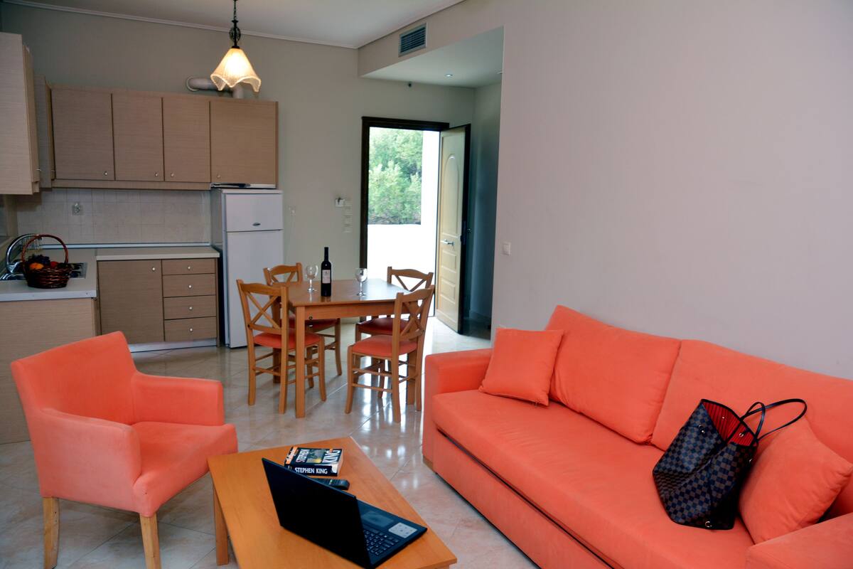 A modern living room with a bright orange sofa, a wooden coffee table with a laptop, and a dining area with wooden chairs and a table.