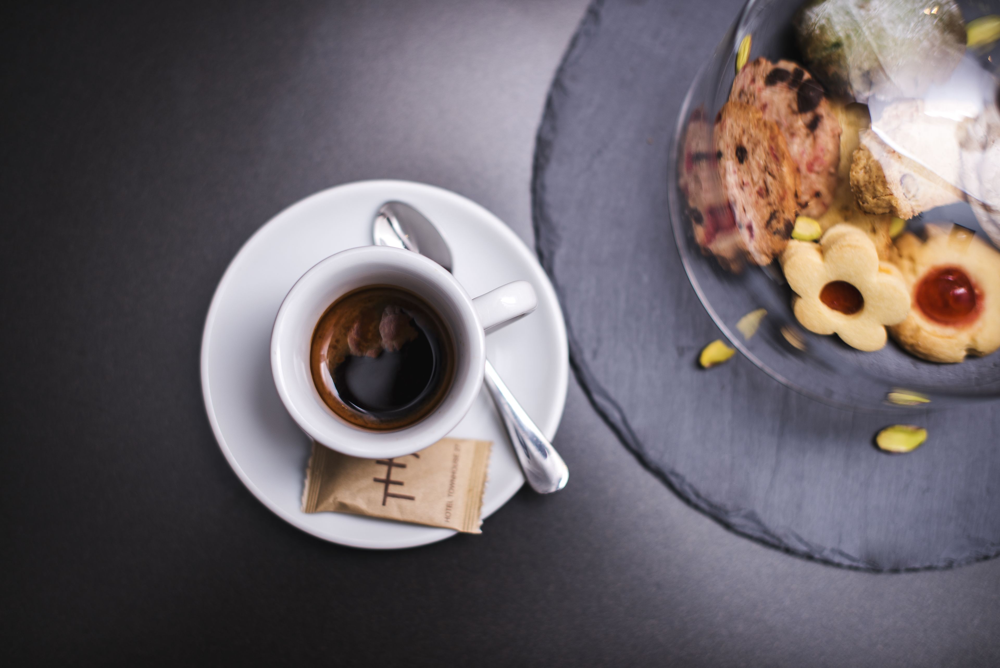 A cup of coffee on a white saucer with a spoon, a small packet of sugar, and a glass containing various pastries and a drink.
