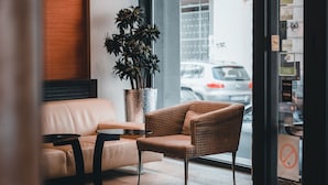 Eine moderne Hotelhalle mit Ledersofa, Korbsessel, Topfpflanze und Glastür zum Außenbereich.