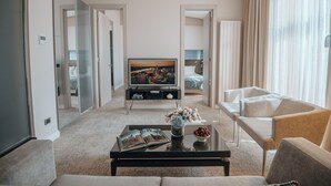Una habitación de hotel moderna con una sala de estar que incluye un sofá, sillones, una mesa de centro con un libro y flores, un televisor y una cama con vistas a un paisaje urbano.