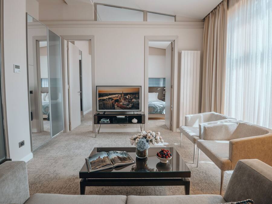 Et moderne hotelværelse med opholdsområde, der indeholder en sofa, lænestole, et sofabord med en bog og blomster, et fjernsyn og en seng med udsigt over bybilledet.