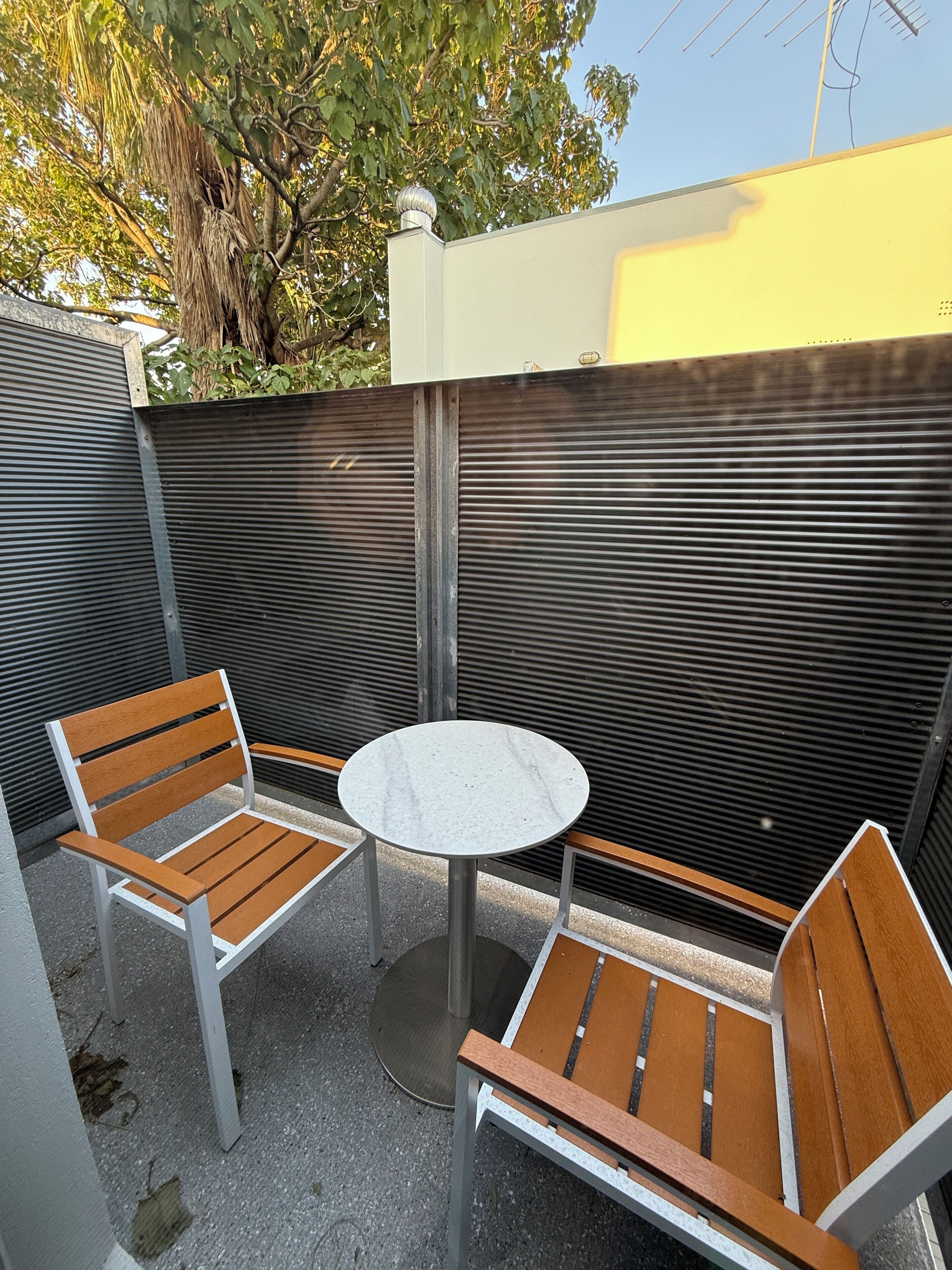 A patio area with two wooden chairs and a small round table, surrounded by a metal fence and a tree.