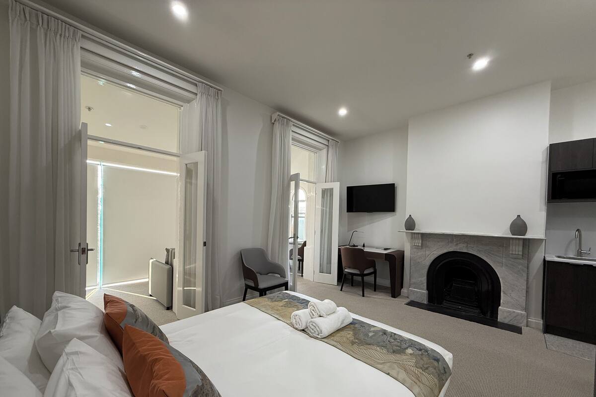 A modern hotel room with a large bed, a desk with a chair, a fireplace, and a TV mounted on the wall.