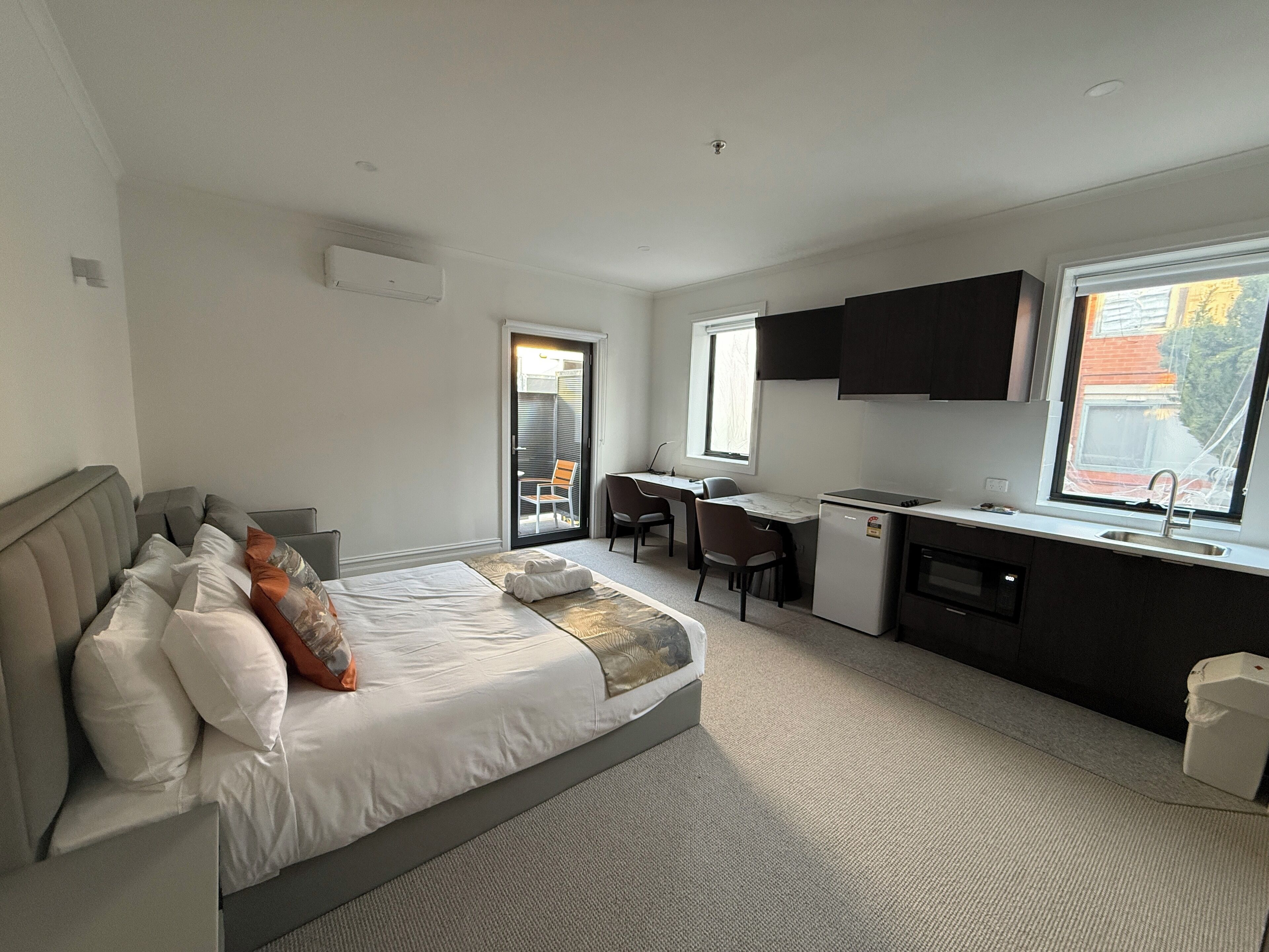 A modern hotel room with a bed, a desk, a kitchenette, and a balcony.