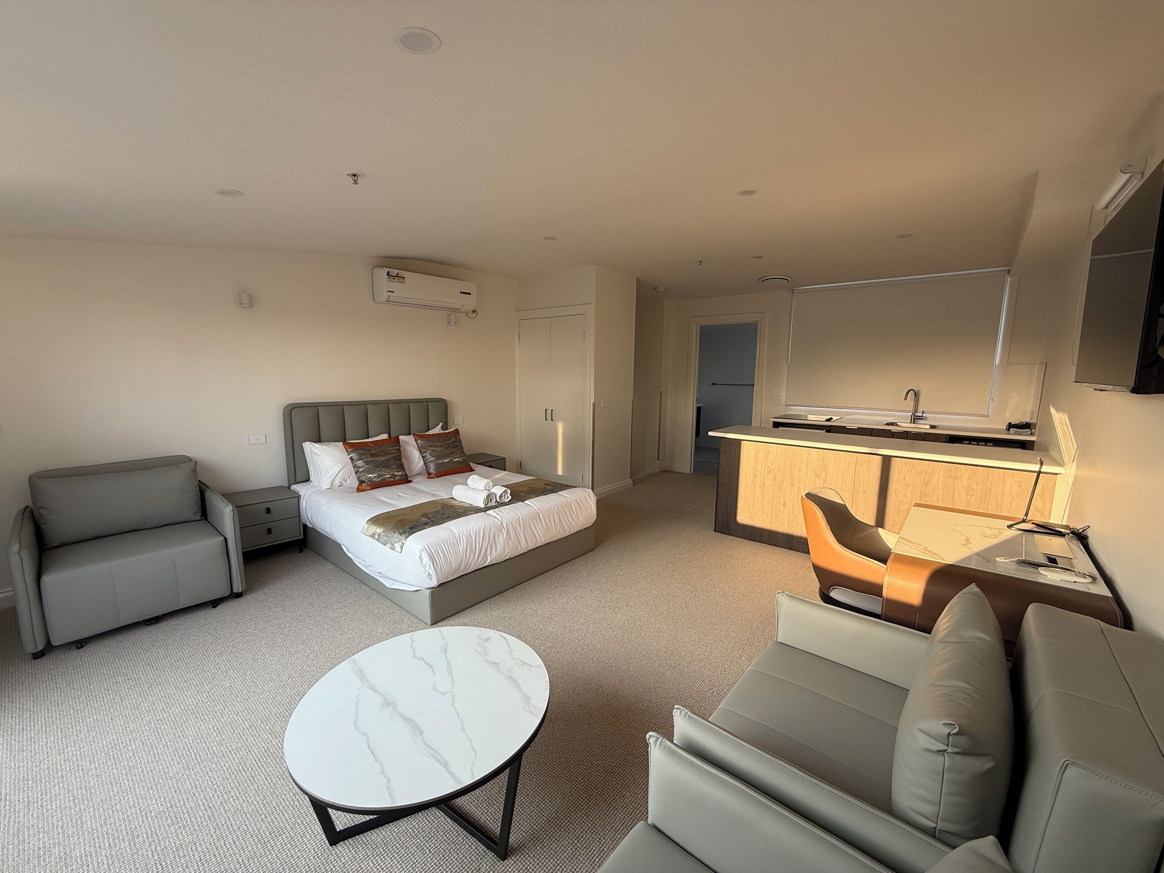 A modern hotel room with a bed, a sofa, a small round table, a desk with a mirror, and a kitchenette.