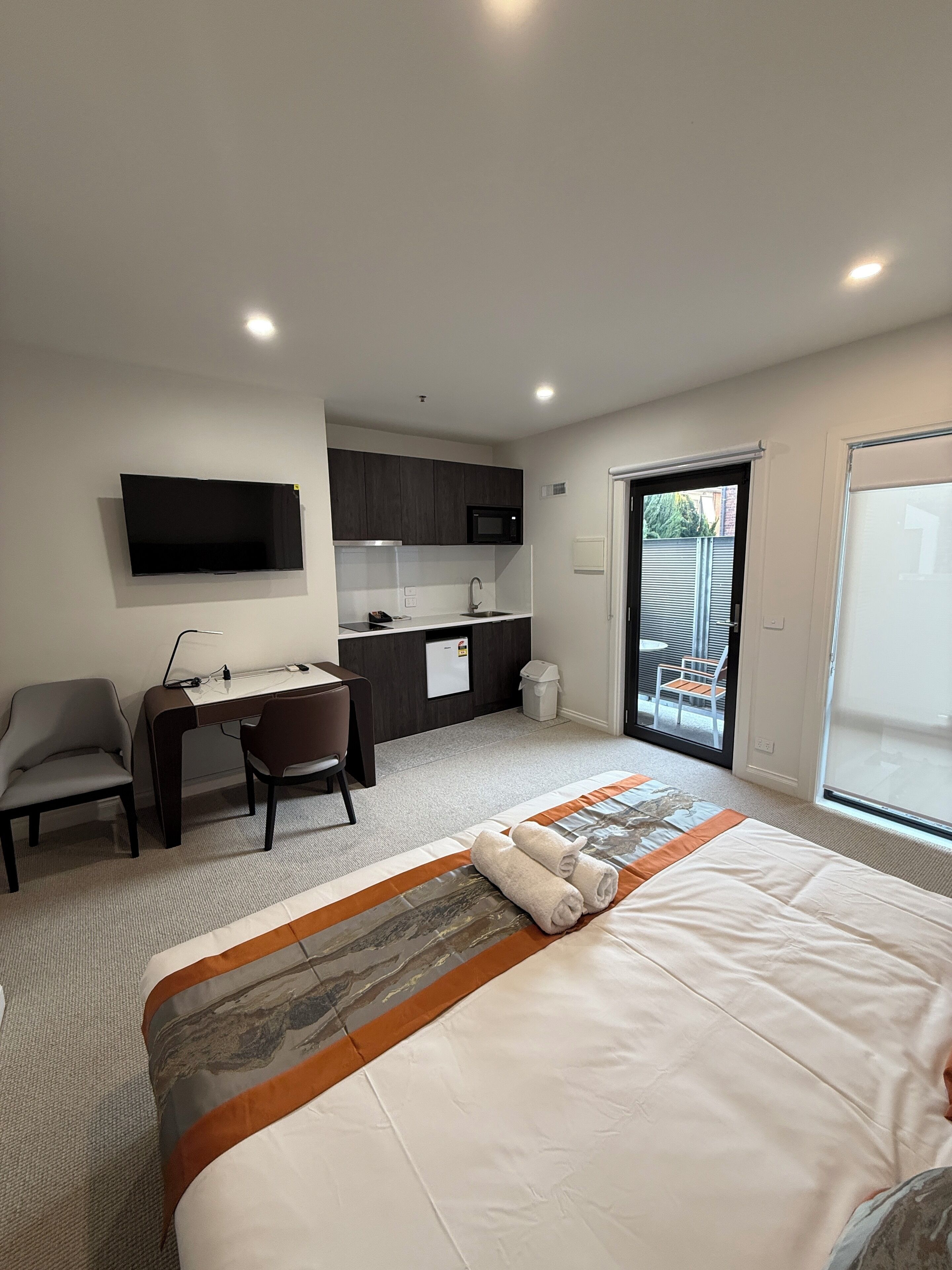 Ein modernes Hotelzimmer mit einem großen Bett, einem Schreibtisch mit Stuhl, einem Flachbildfernseher, einer Küchenzeile und einer Glastür zum Außenbereich.