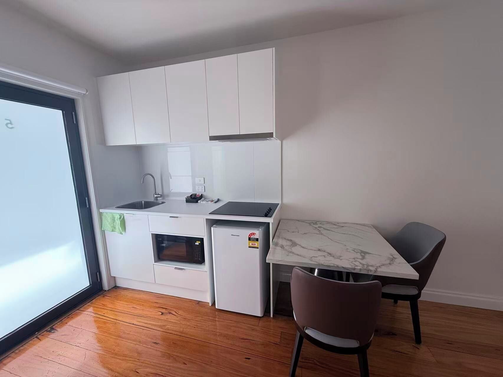 Cocina compacta con armarios blancos, horno integrado, microondas y un refrigerador pequeño. Hay una mesa de comedor con dos sillas y vista al exterior por una puerta de vidrio.