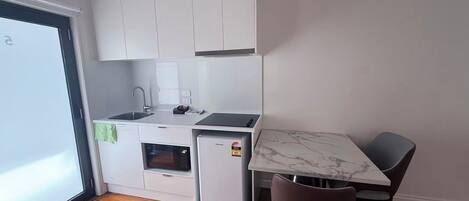 Cocina compacta con armarios blancos, horno integrado, microondas y un refrigerador pequeño. Hay una mesa de comedor con dos sillas y vista al exterior por una puerta de vidrio.