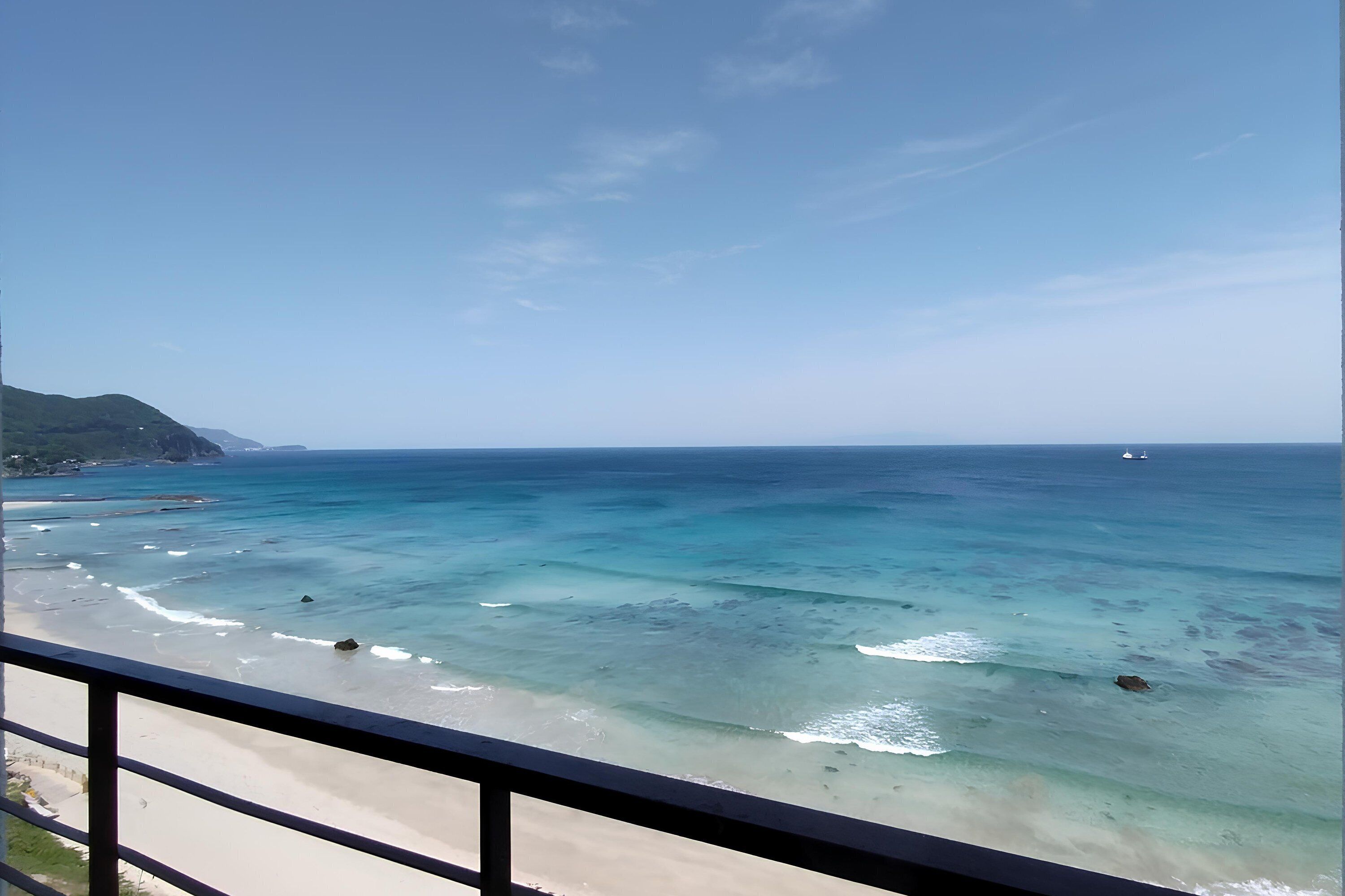 Vista costera desde un balcón con mar azul transparente, playa de arena y montañas a lo lejos.