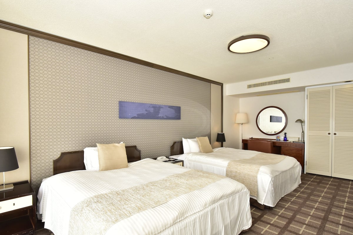 A hotel room with two beds, a nightstand, a desk, a chair, a mirror, and a wardrobe.