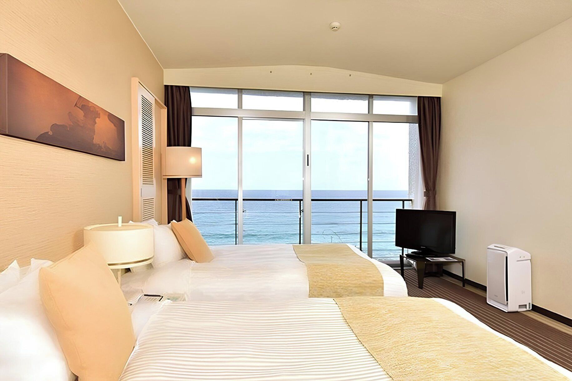 A hotel room with two beds, a TV, a heater, and a large window with a view of the sea.
