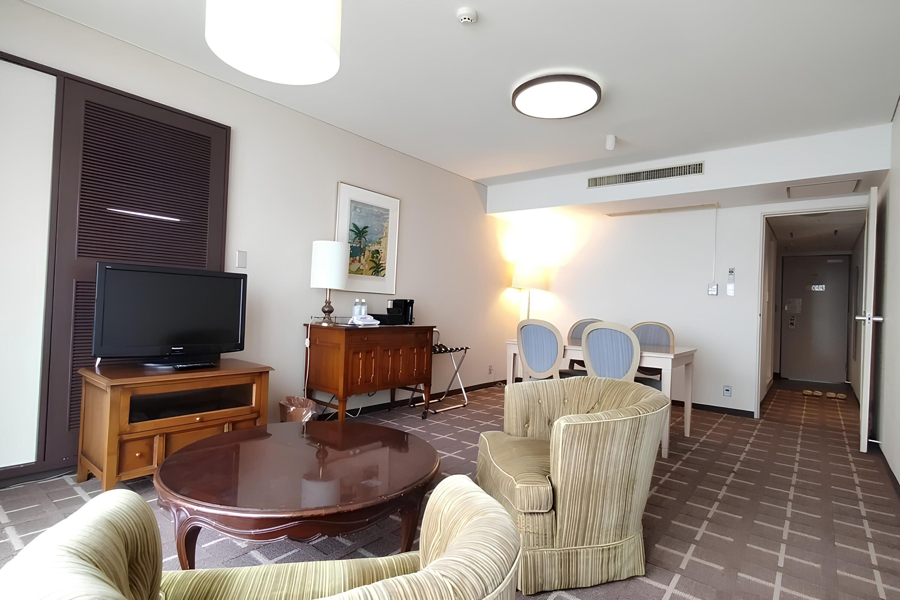A hotel room with a television on a wooden stand, a dining table with chairs, a sofa, a lamp, and a painting on the wall.