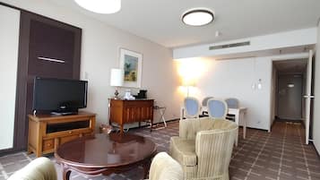 A hotel room with a television on a wooden stand, a dining table with chairs, a sofa, a lamp, and a painting on the wall.