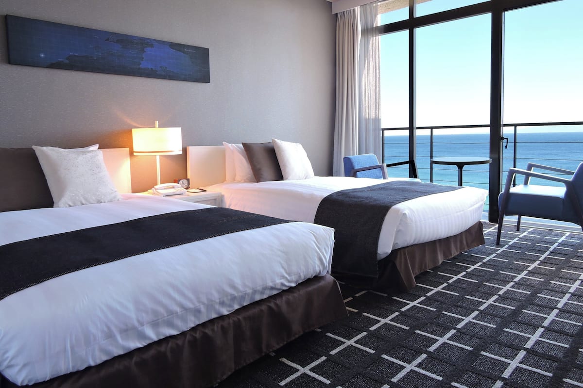 A hotel room with two beds, a nightstand, a lamp, a chair, and a large window with a view of the ocean.