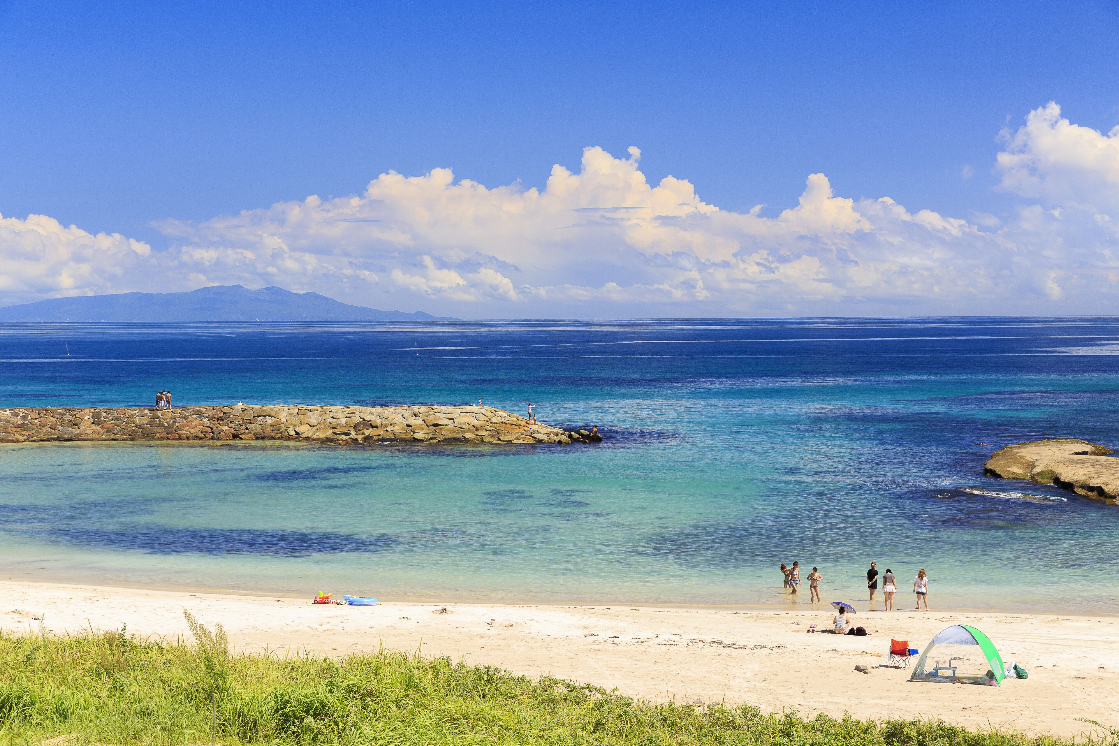 A beach with clear blue water, a rocky outcrop, and a few people.