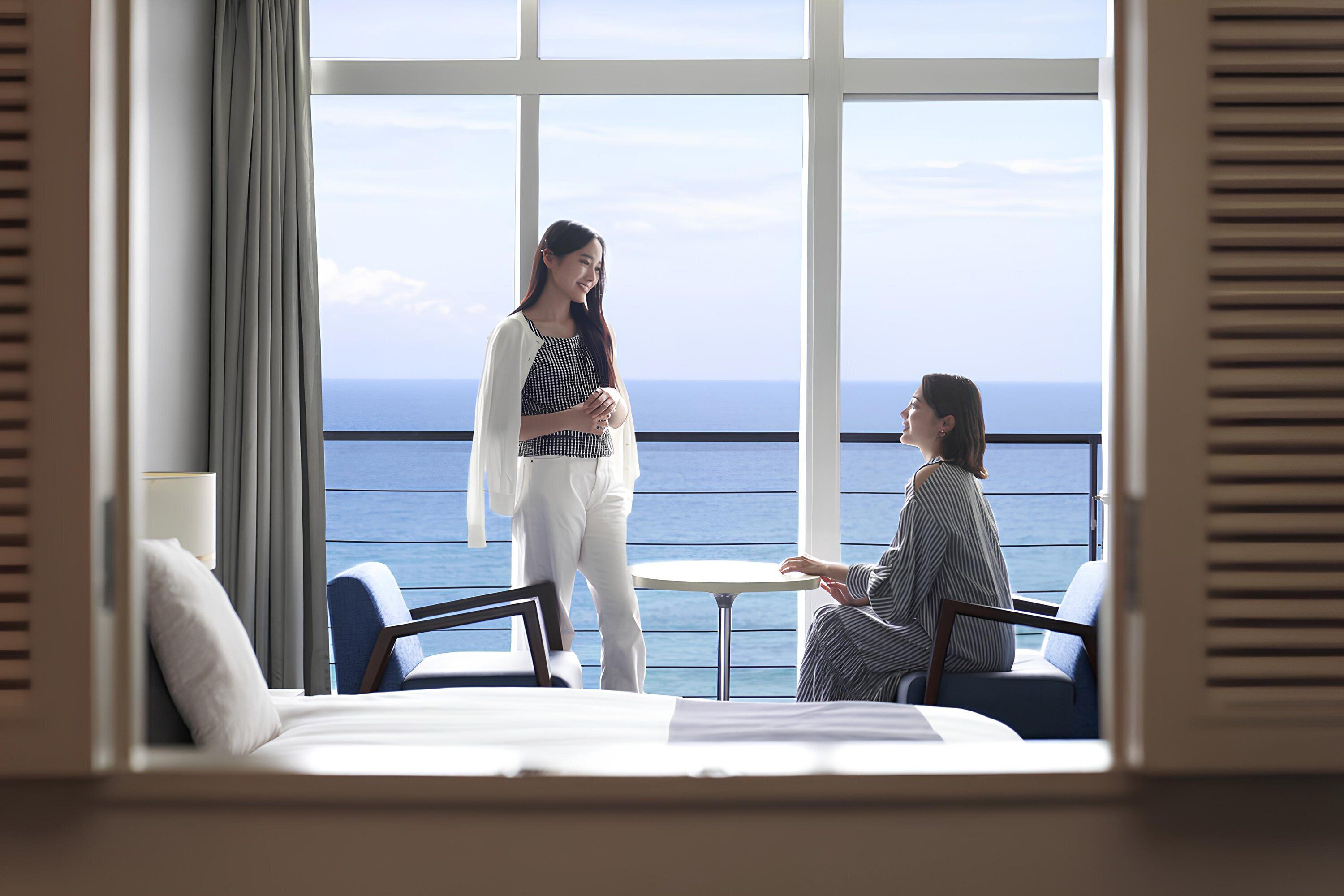 A room with a large window overlooking the sea, a bed, a small round table, and two chairs.