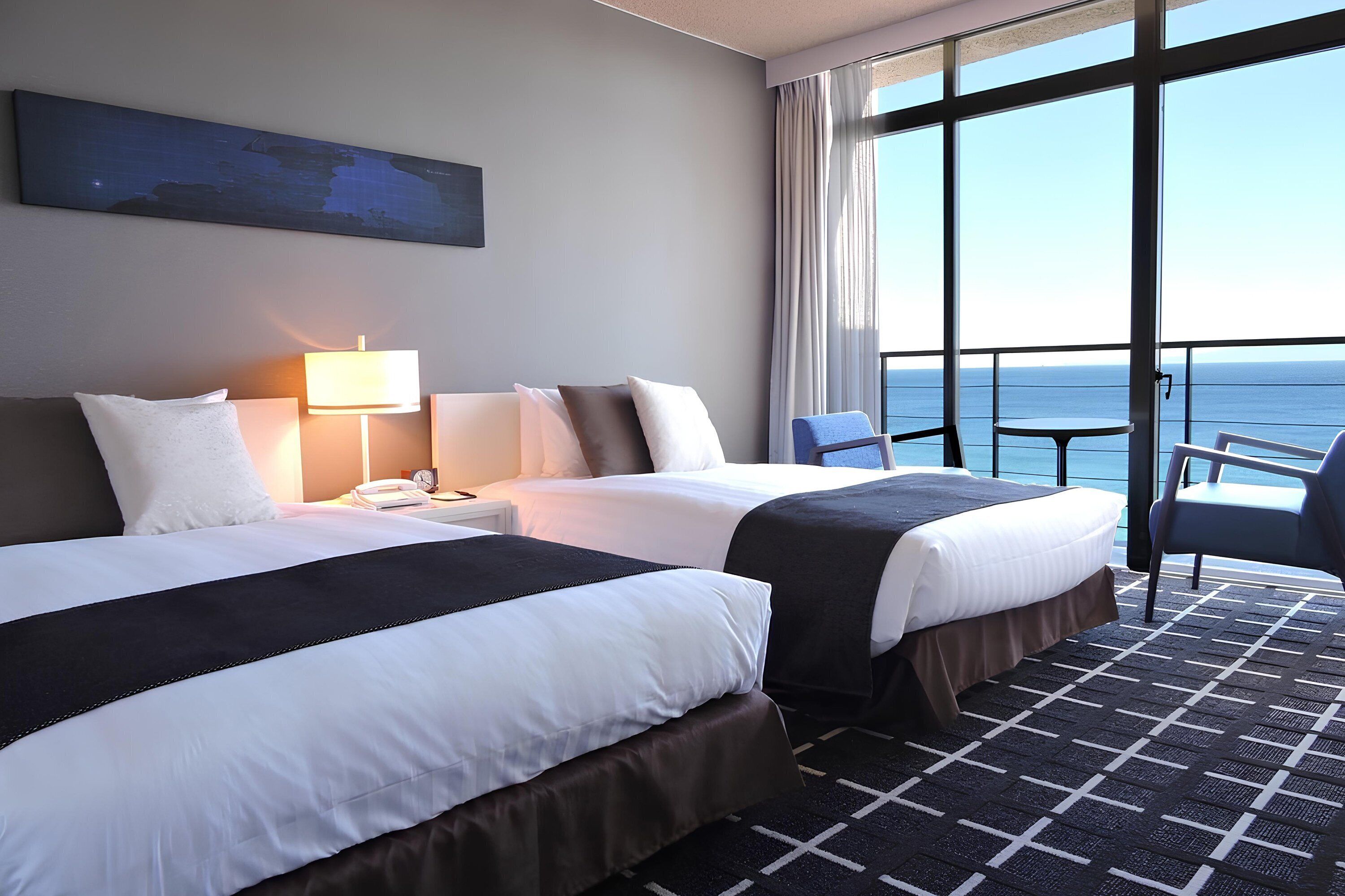 A hotel room with two beds, a desk with a lamp, a chair, and a view of the sea through large windows.