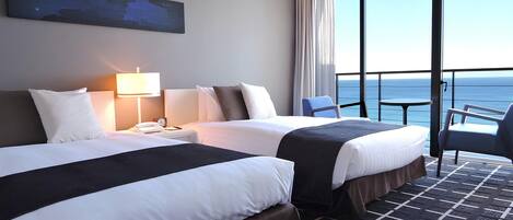 A hotel room with two beds, a desk with a lamp, a chair, and a view of the sea through large windows.