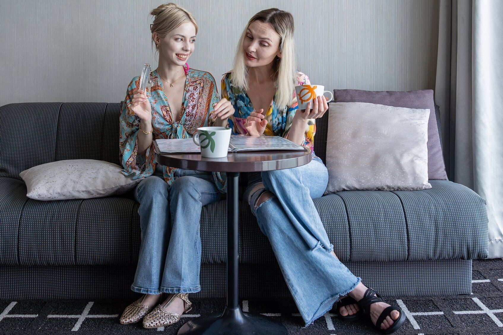 Two people sitting on a grey sofa with a small round table between them, holding a mug and a glass.