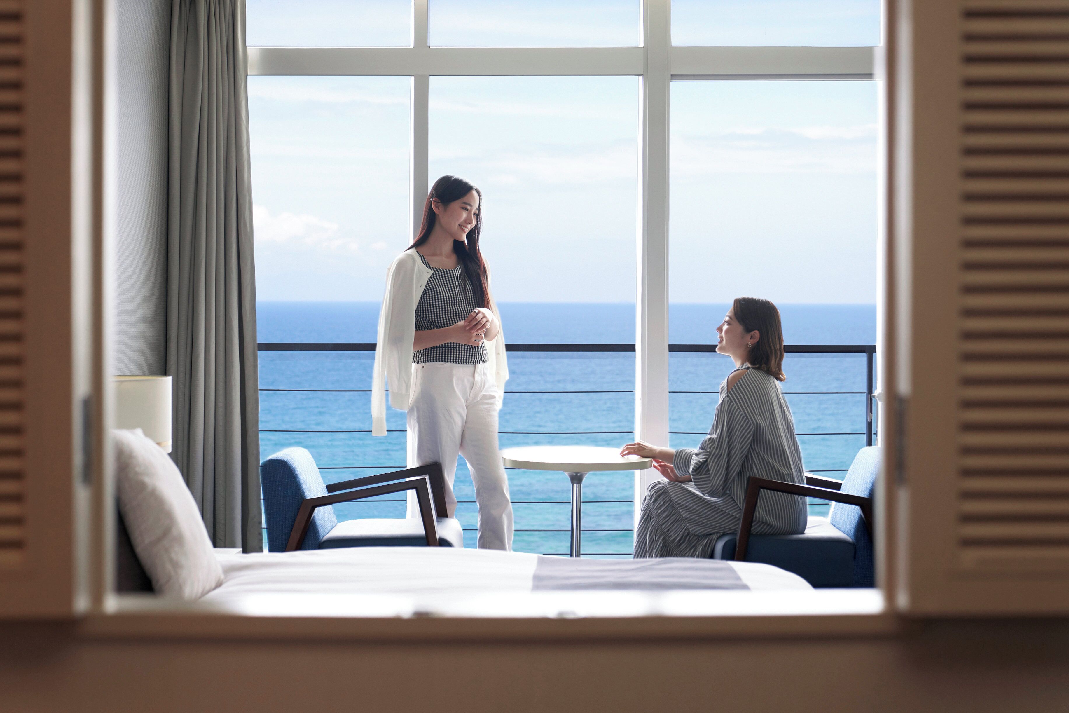 Une pièce dotée d’une grande fenêtre donnant sur la mer, d’un lit, d’une petite table et de deux chaises.