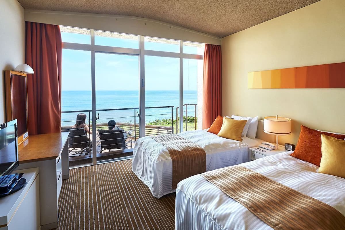 A hotel room with two beds, a desk with a computer, and a view of the ocean through large windows.