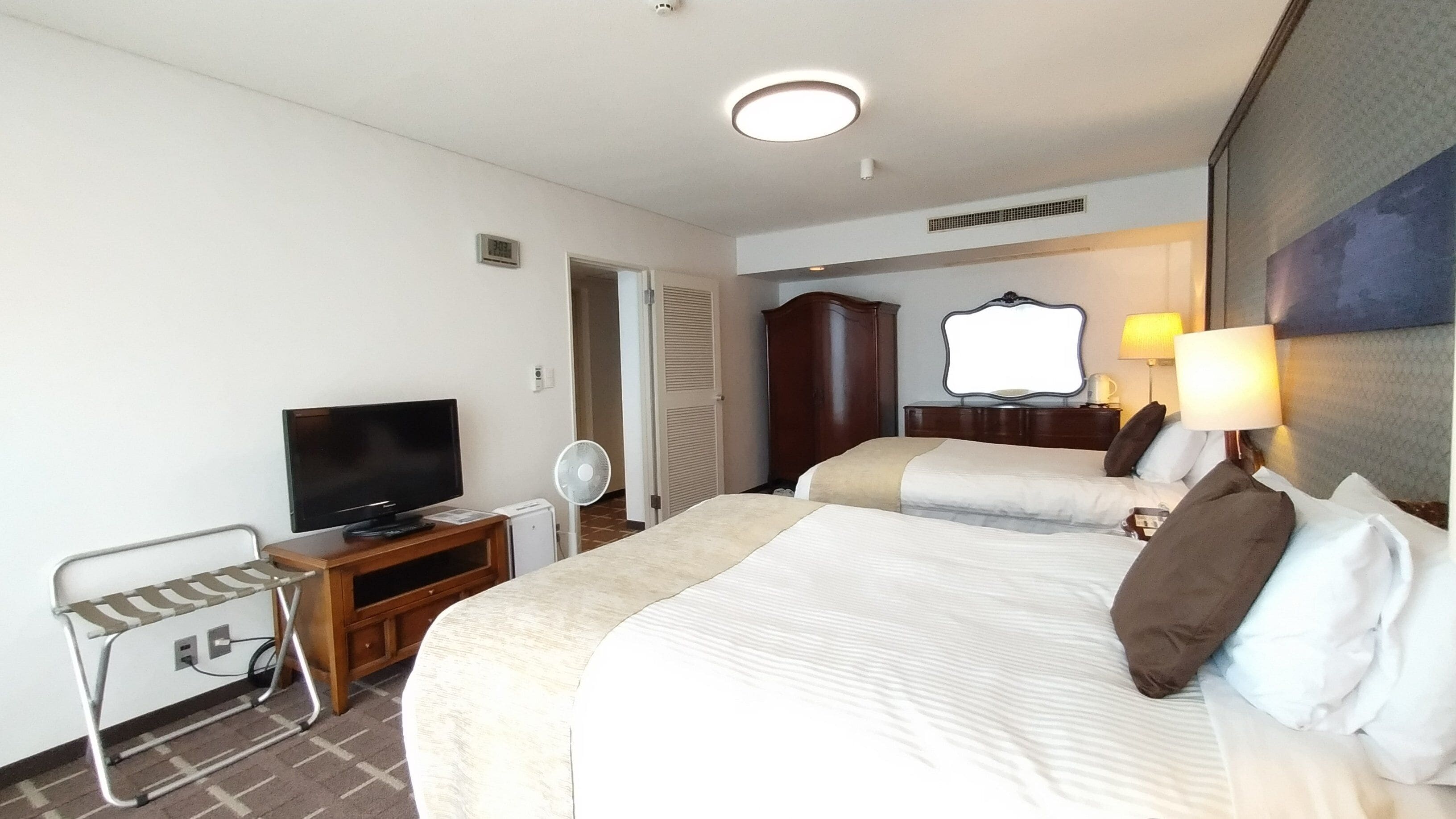 A hotel room with two beds, a TV, a desk, a chair, a fan, and a lamp.