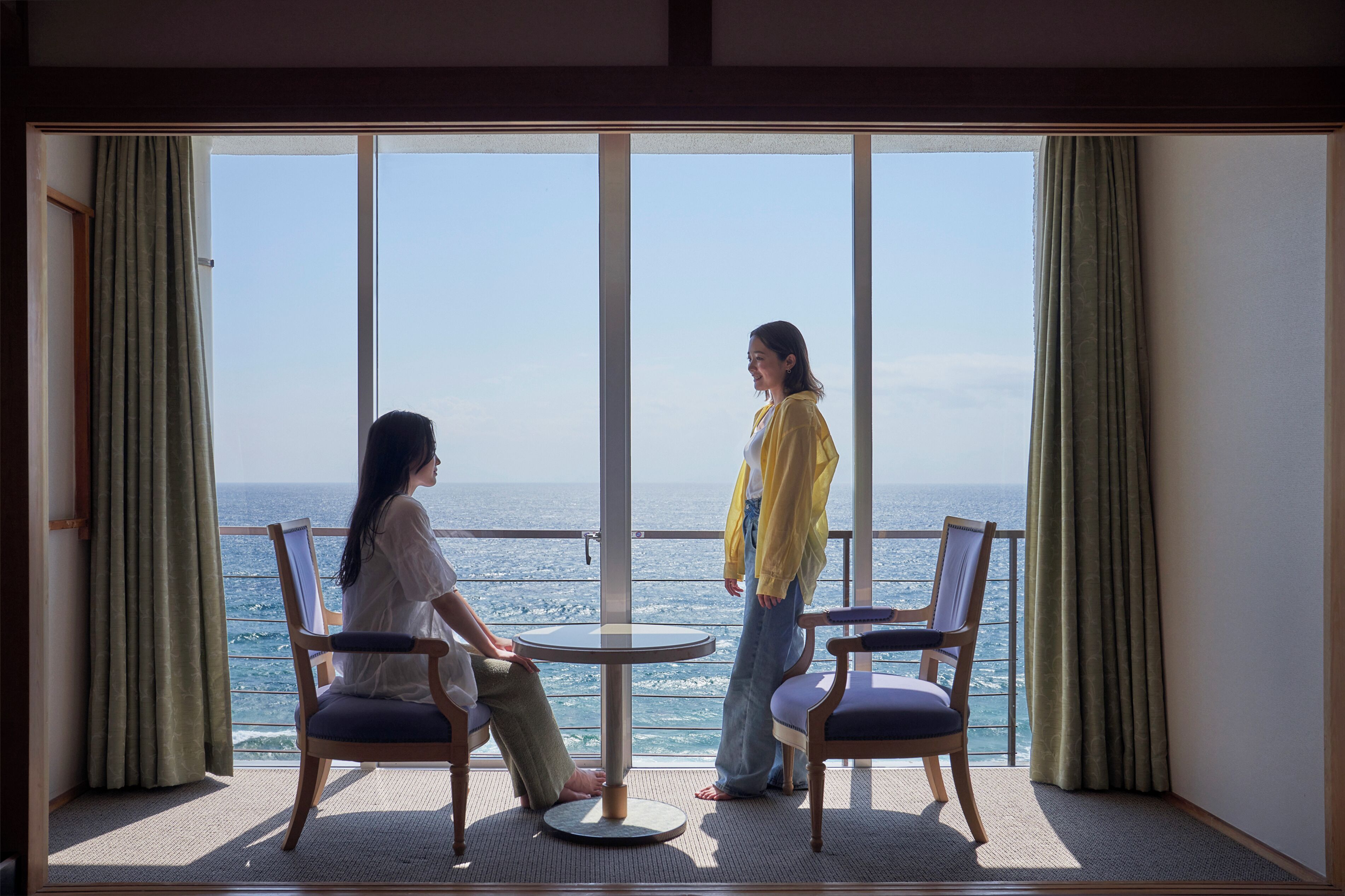 A room with large windows overlooking the sea, two chairs, and a small round table.