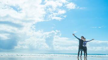 Two people standing on a beach with their arms outstretched, facing the ocean under a partly cloudy sky.