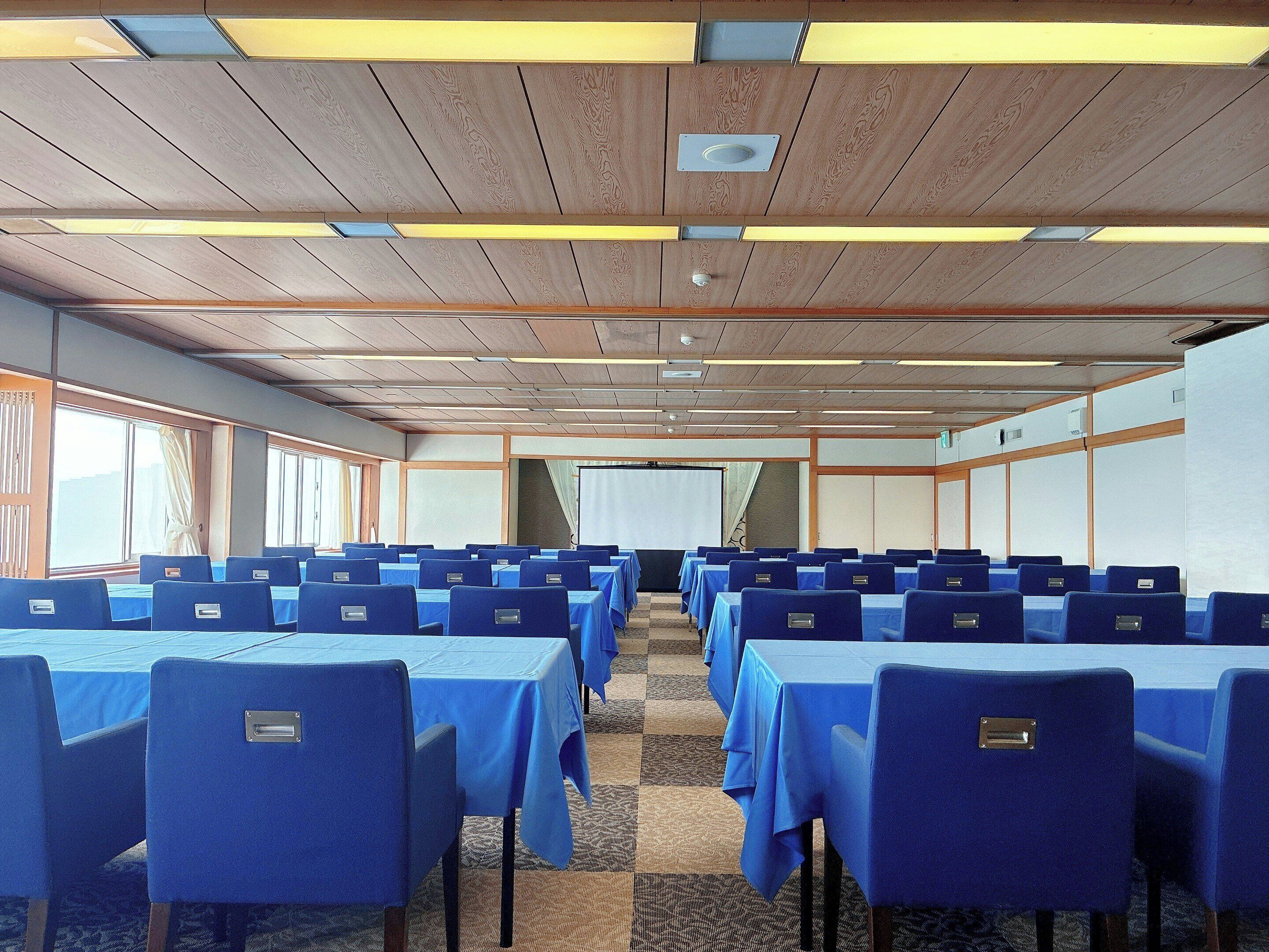 Un salón de conferencias con filas de mesas y sillas, paneles de techo de madera y ventanas amplias.
