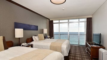 A hotel room with two beds, a TV, a desk, and a large window with a view of the sea.