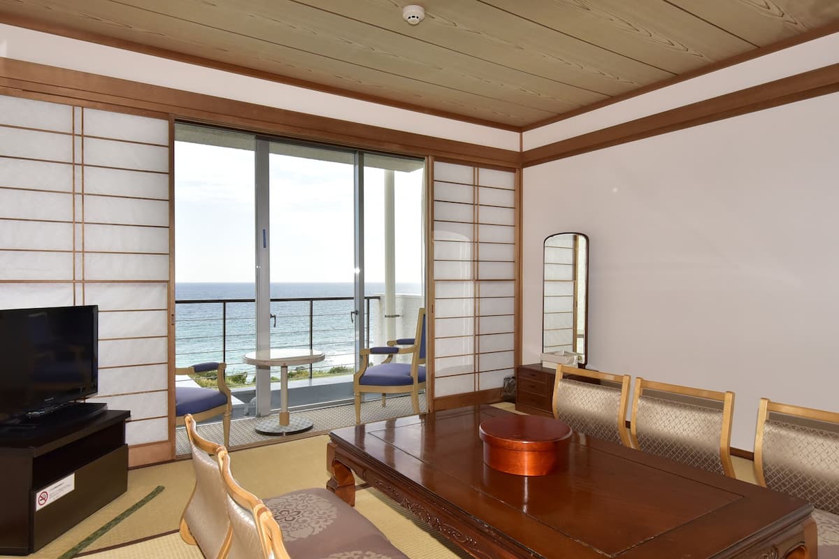 A room with a wooden ceiling, a large sliding glass door leading to a balcony with a view of the sea, a dining table with chairs, a television, and a cabinet.