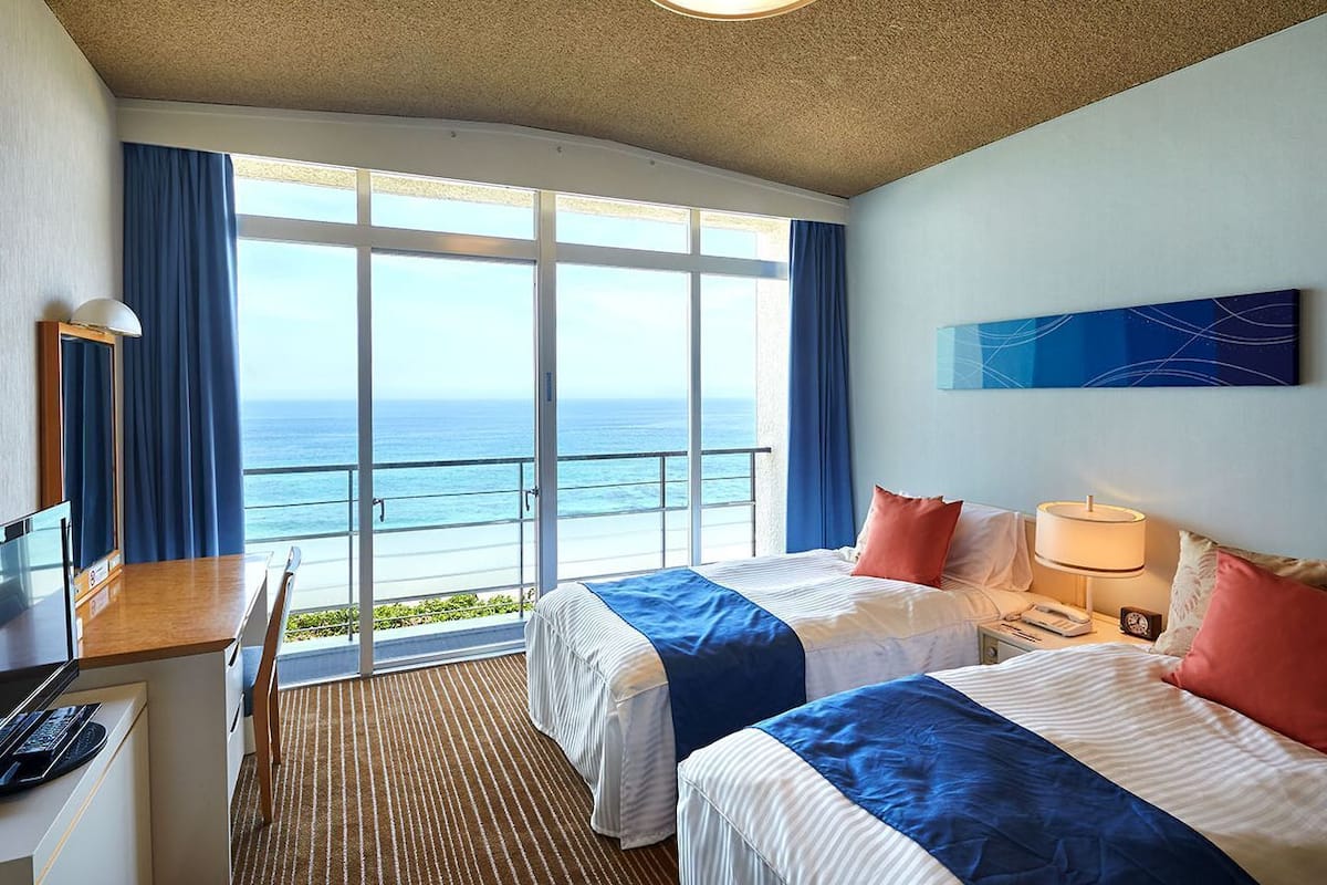 A hotel room with two beds, a desk, a chair, a lamp, and a view of the ocean.