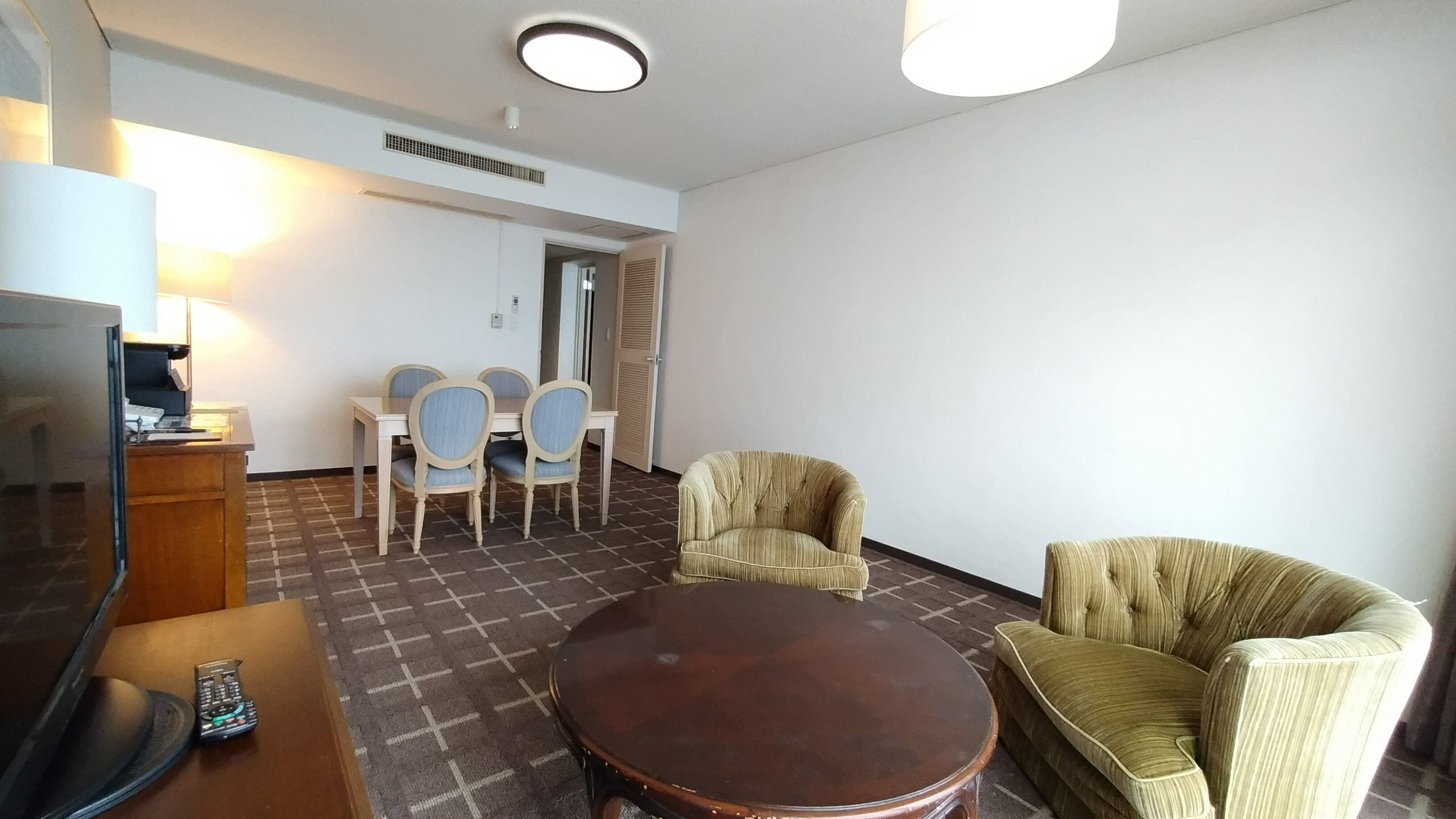 Une chambre d’hôtel équipée d’une télévision, d’une table en bois, d’une petite table ronde, de deux fauteuils et d’un coin repas avec une table et des chaises.