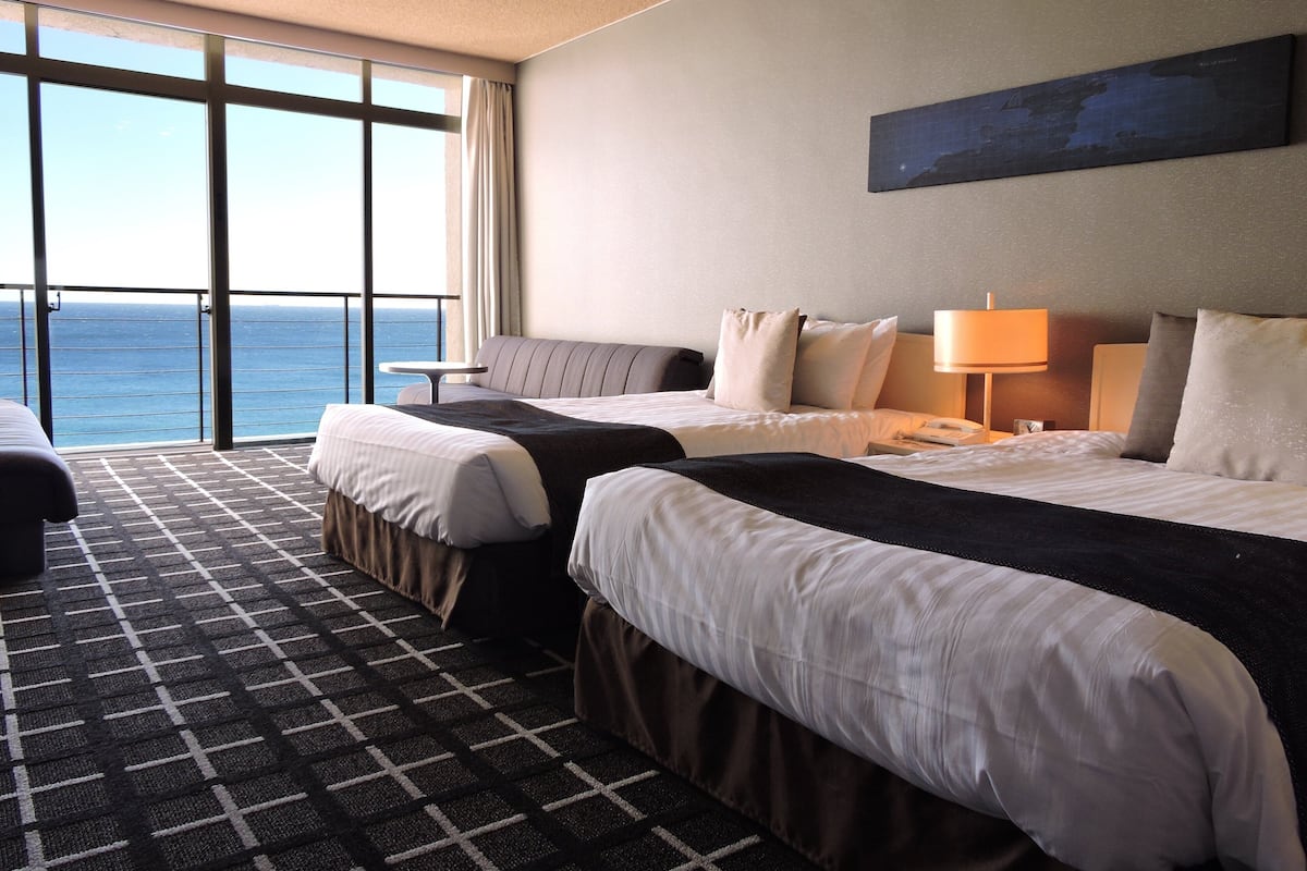 A hotel room with two beds, a view of the ocean, a large window, and a painting on the wall.